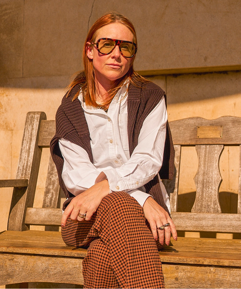 Eddie wearing a white shirt and patterned trousers sitting on a wooden bench in a sunlit stone courtyard.