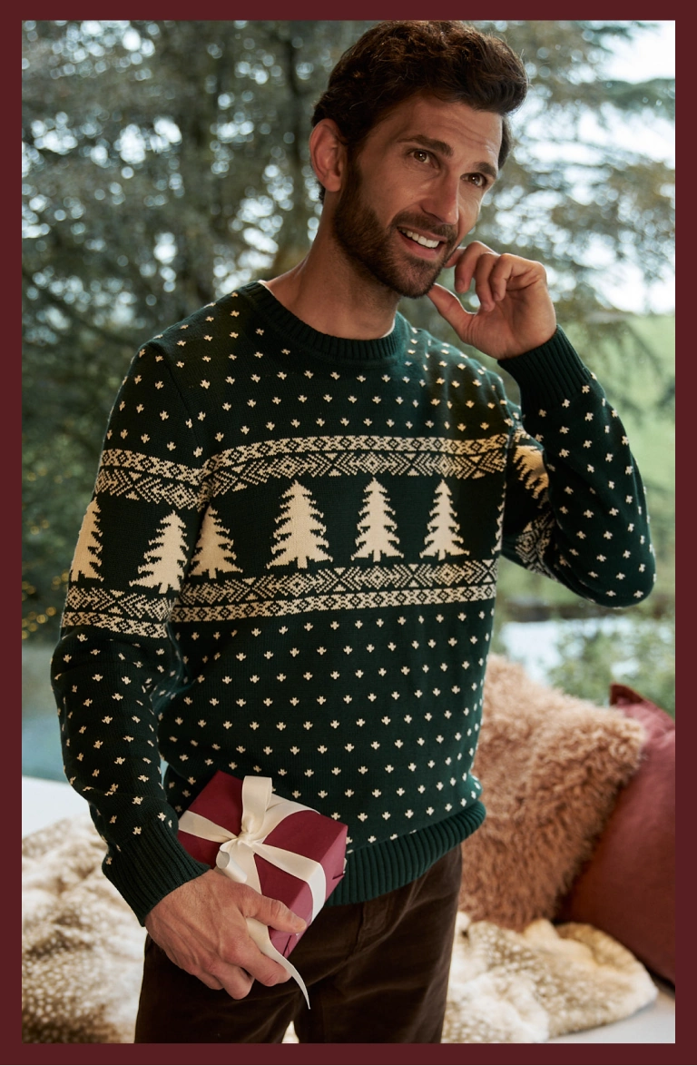 Man wearing a dark green Christmas jumper with a tree pattern, holding a gift with a 50% off graphic