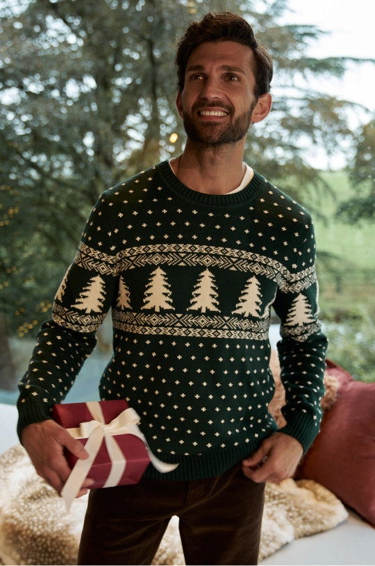 Man wearing a dark green Christmas jumper with a tree pattern, holding a wrapped gift